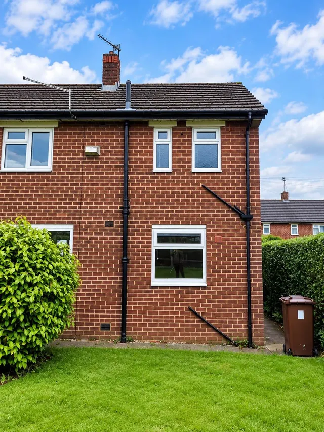The gable end of a red-brick semi-detached house, with a chimney, soil pipe and two small windows.